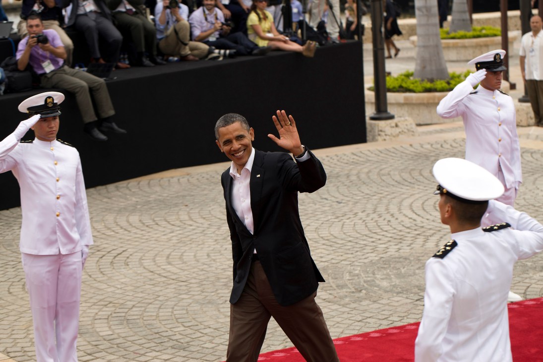 Barack Obama waves while walking on red carpet flanked by Colombian naval officers in white uniforms.