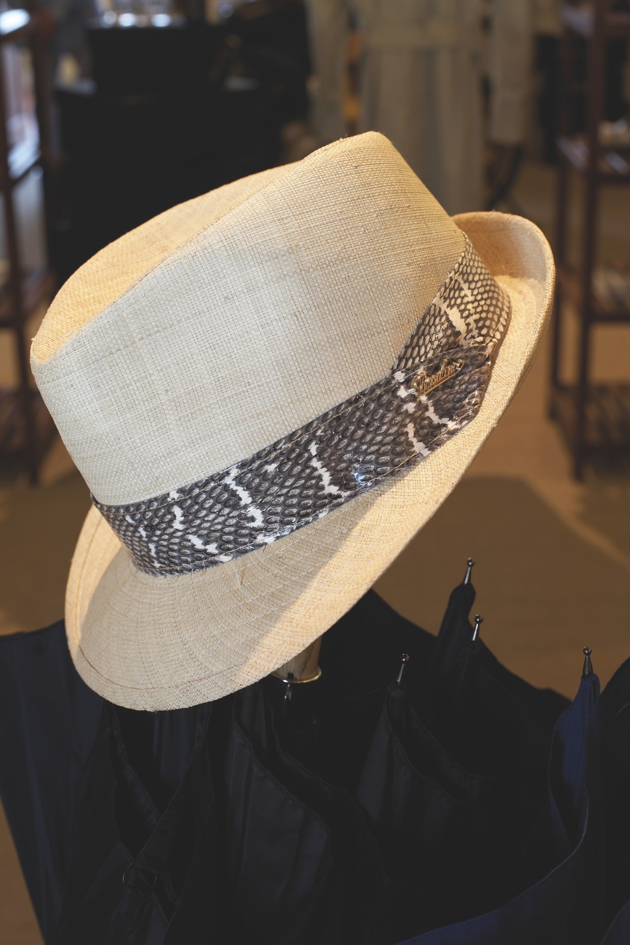 Cream-colored straw fedora with black patterned feather band displayed on a stand.