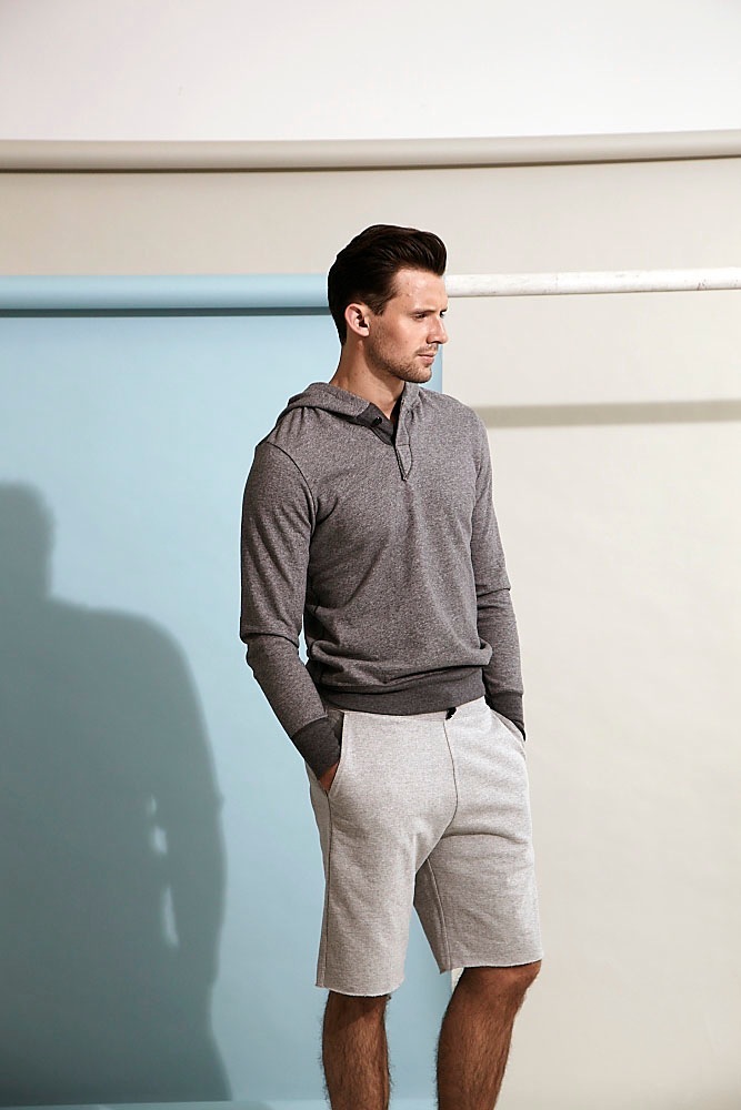 Man wearing a grey hooded henley and light beige shorts against a minimalist wall.