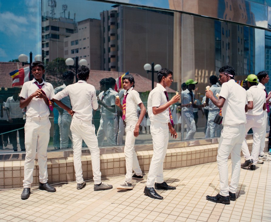 Young men in white clothing eat ice cream in front of a modern building with reflective glass.