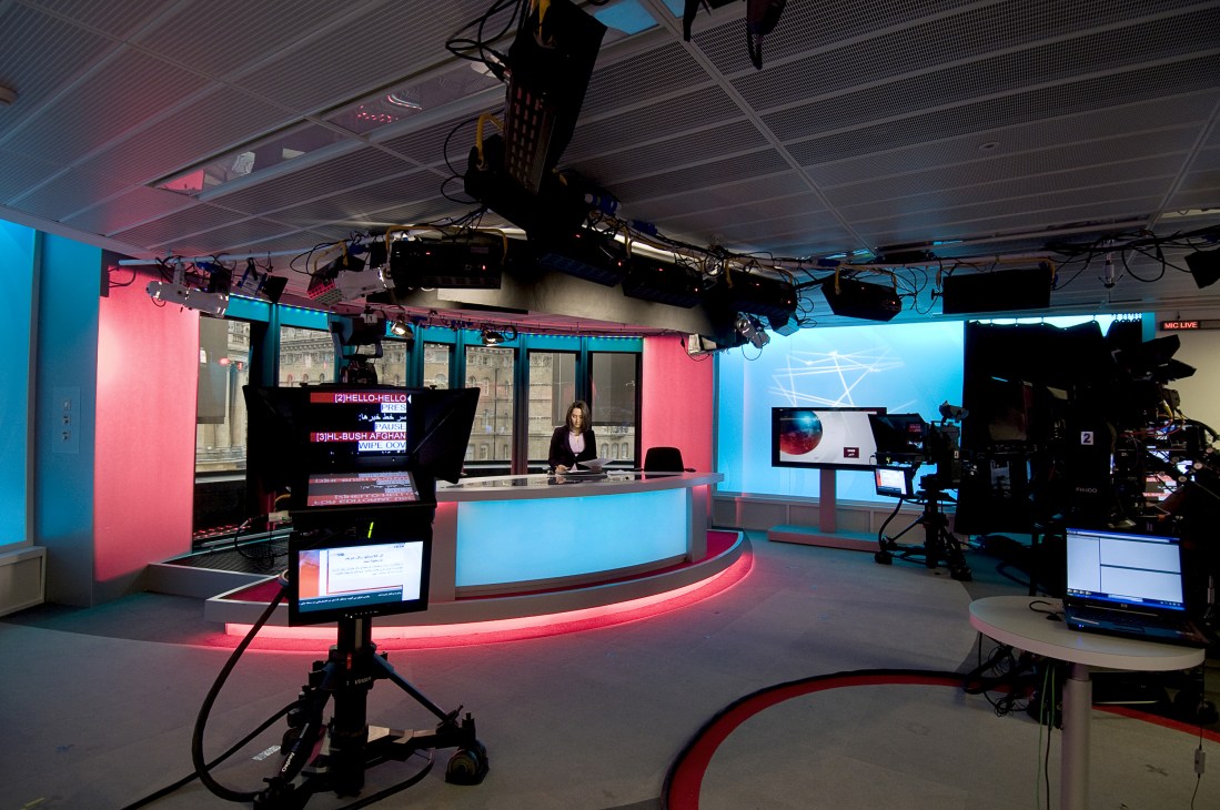 BBC Persian TV news studio with female presenter at curved desk and production equipment with red and blue lighting.