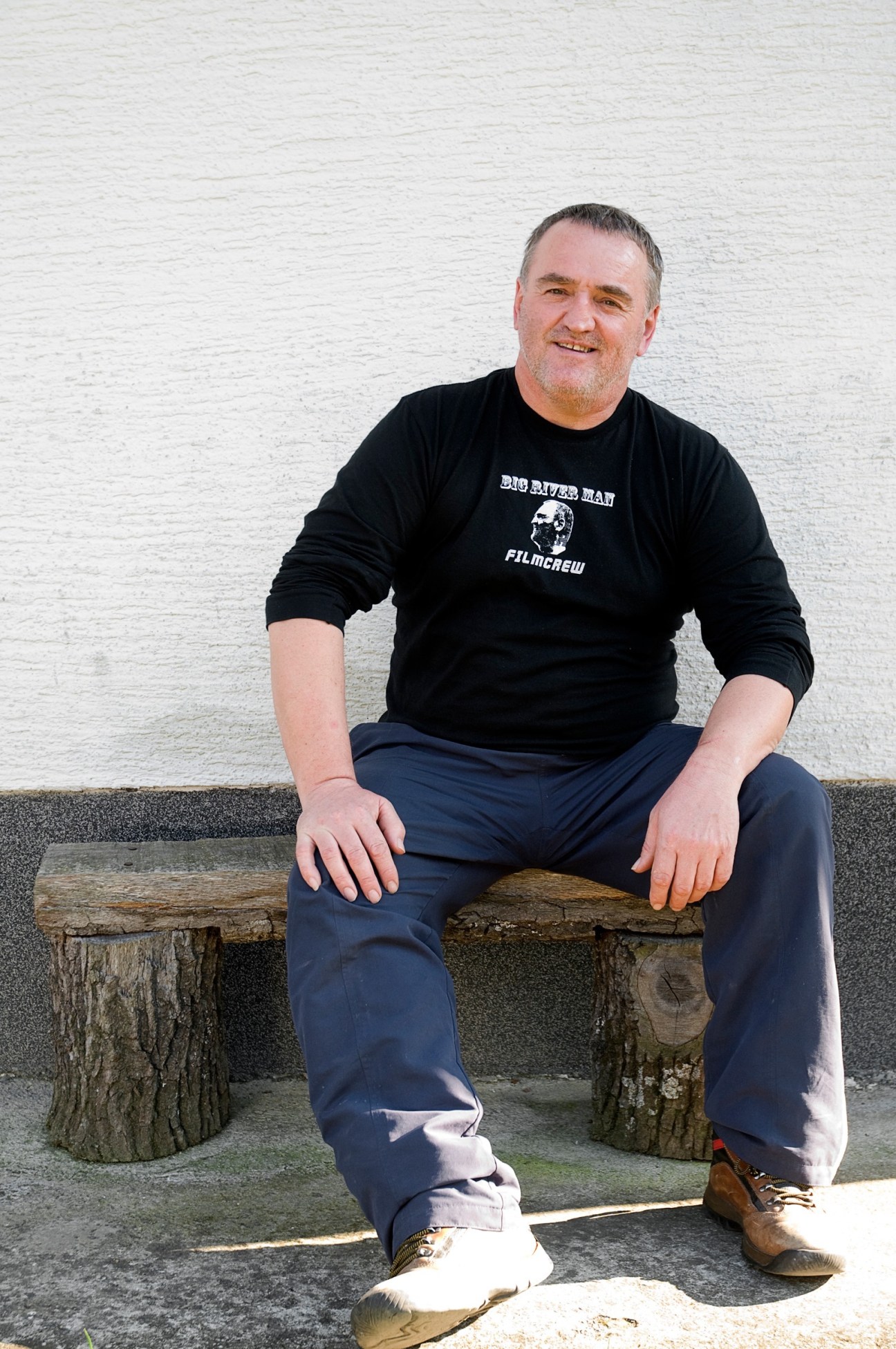 Martin Strel sits on a wooden bench outside a white-walled cottage, wearing a black Big River Man film crew shirt.
