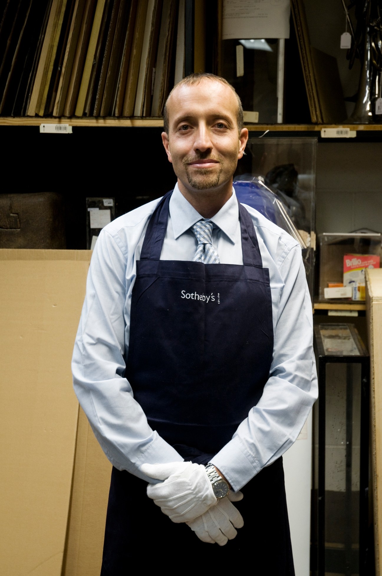 Dominic Cacioppo, wearing a Sotheby's apron over a shirt and tie, stands in a storage room with paintings stacked behind him.