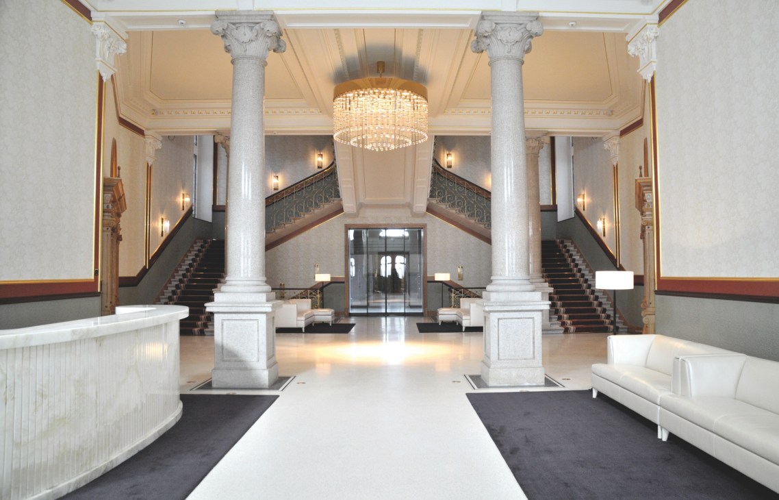 The main lobby and entrance of Dolder Grand featuring symmetrical marble columns, grand staircase, and crystal chandelier.
