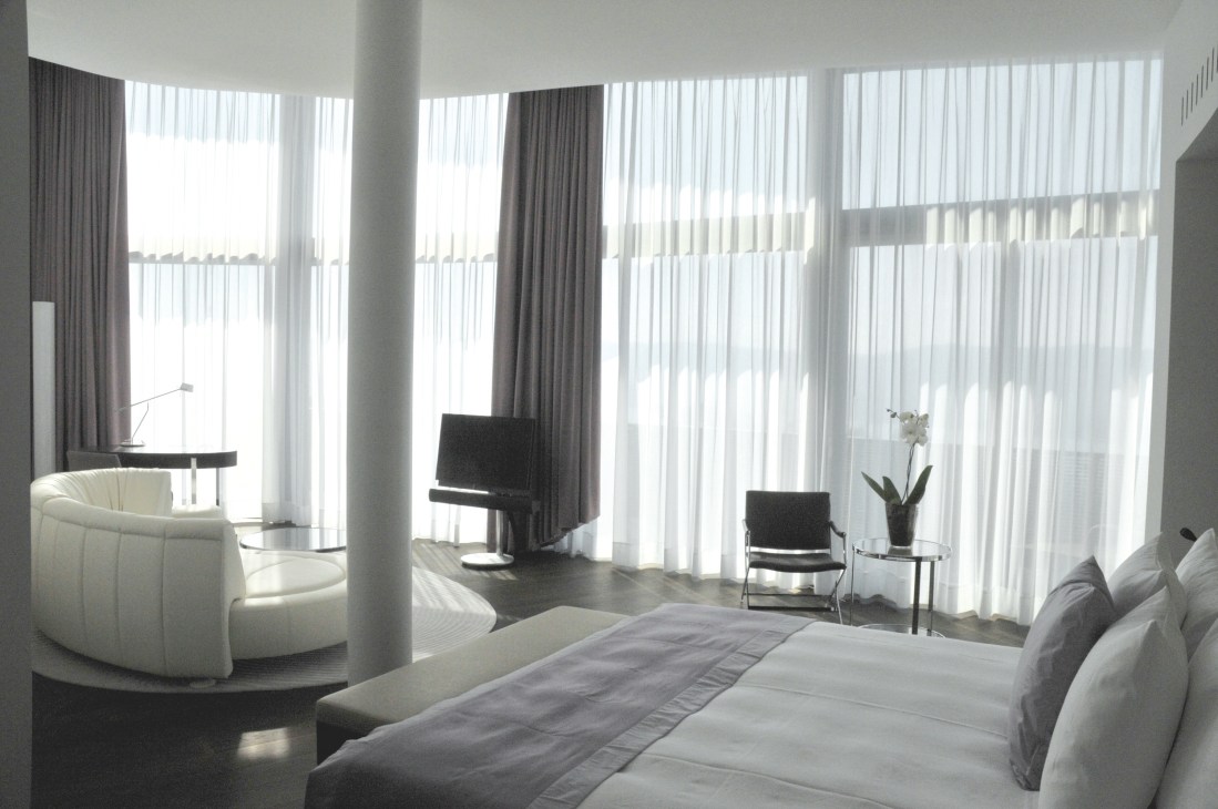 Modern hotel junior suite with floor-to-ceiling windows, white curved seating, and minimalist furnishings at the Dolder Grand.