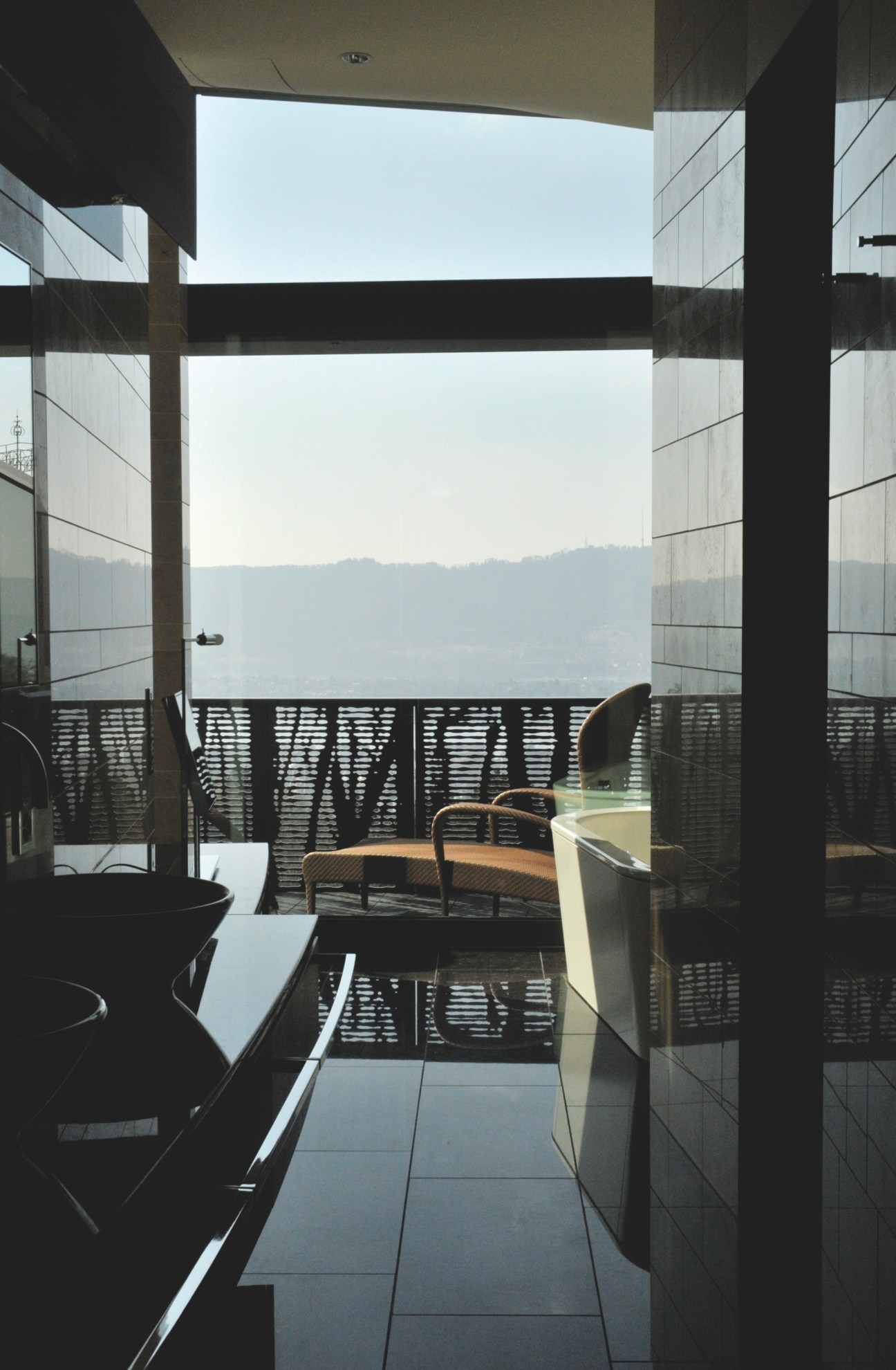 View from a bathroom with bathtub overlooking Zürich and forested hills through floor-to-ceiling windows.