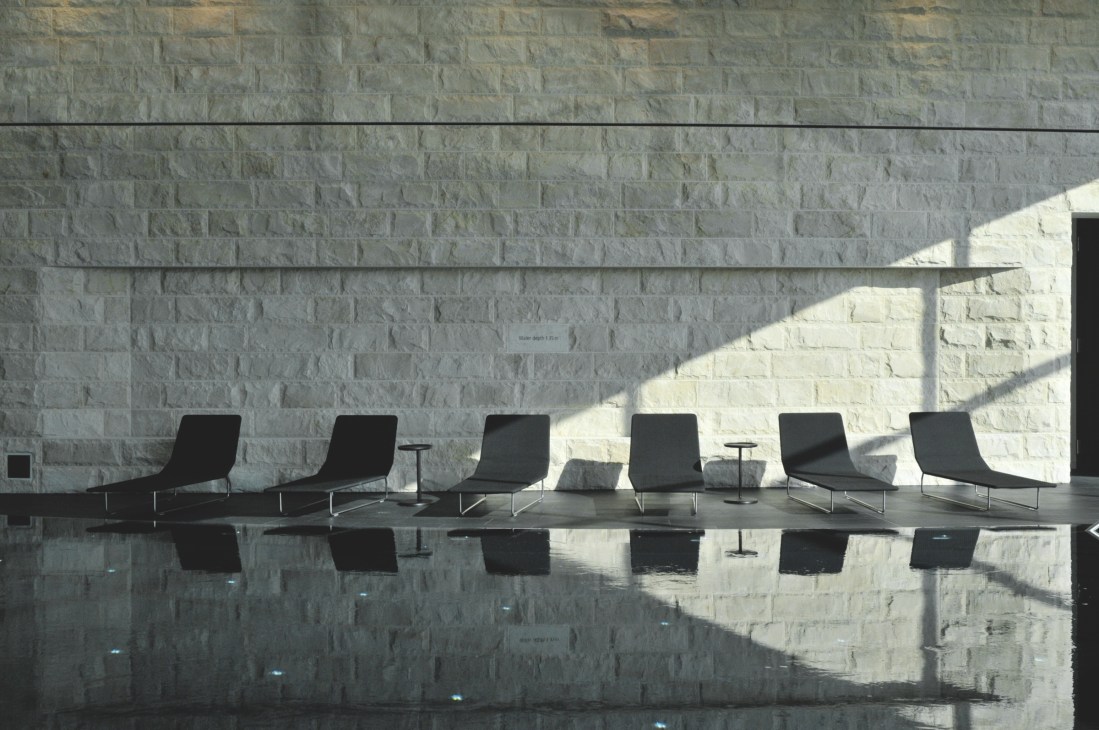 Modern black lounge chairs reflected in still pool water against stone wall at Dolder Grand.