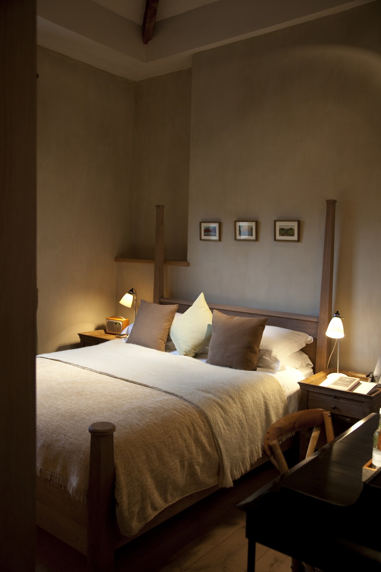 A minimalist hotel bedroom with a wooden four-poster bed, neutral bedding, warm lighting, and framed artwork on pale walls.