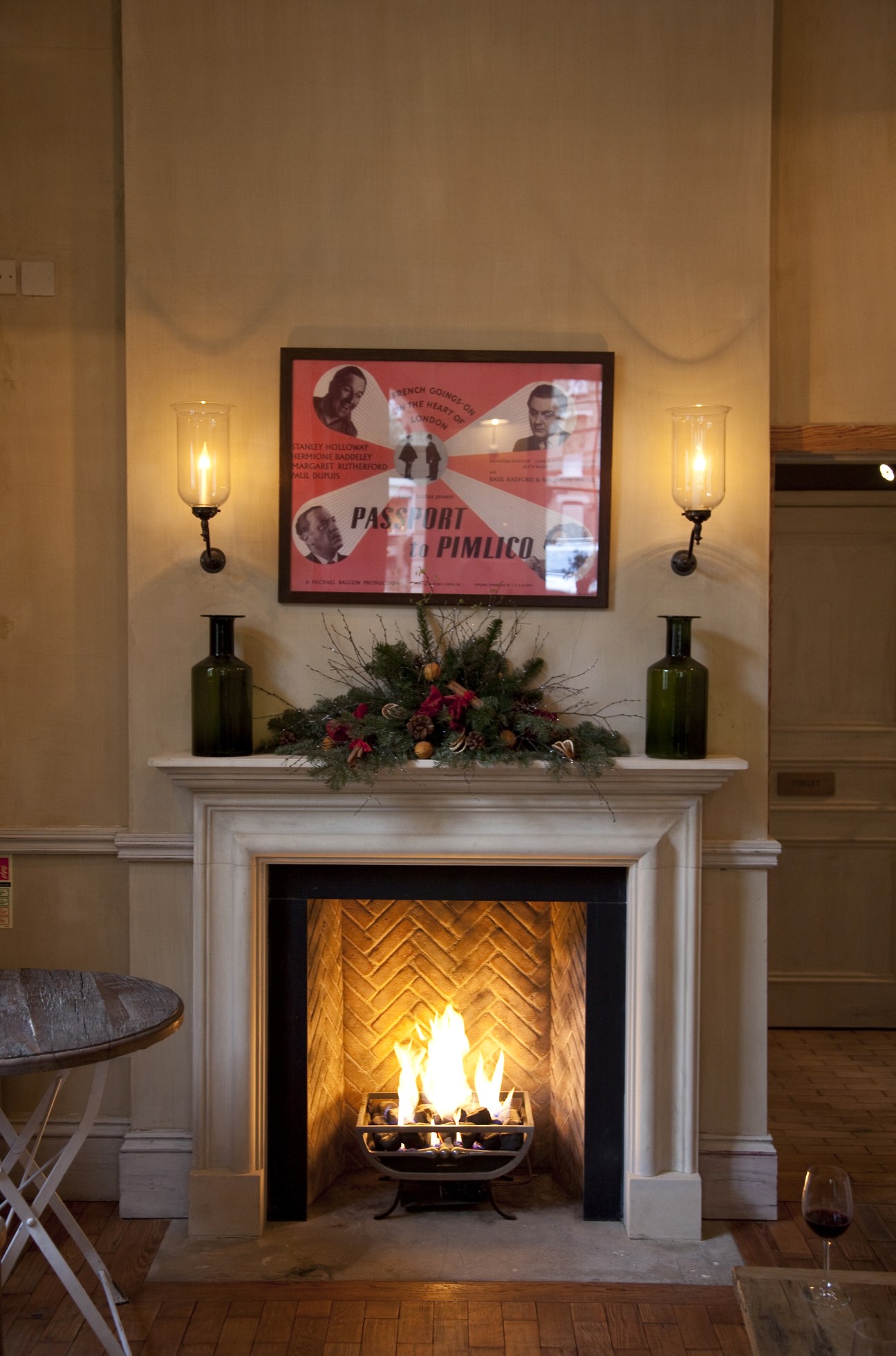 Fireplace with lit fire, mantelpiece decorated with green bottles and holiday garland, vintage film poster above.