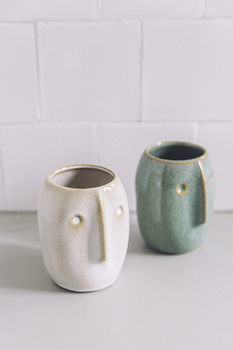 Two ceramic cups with face-like designs, one white and one green, sitting on a light surface