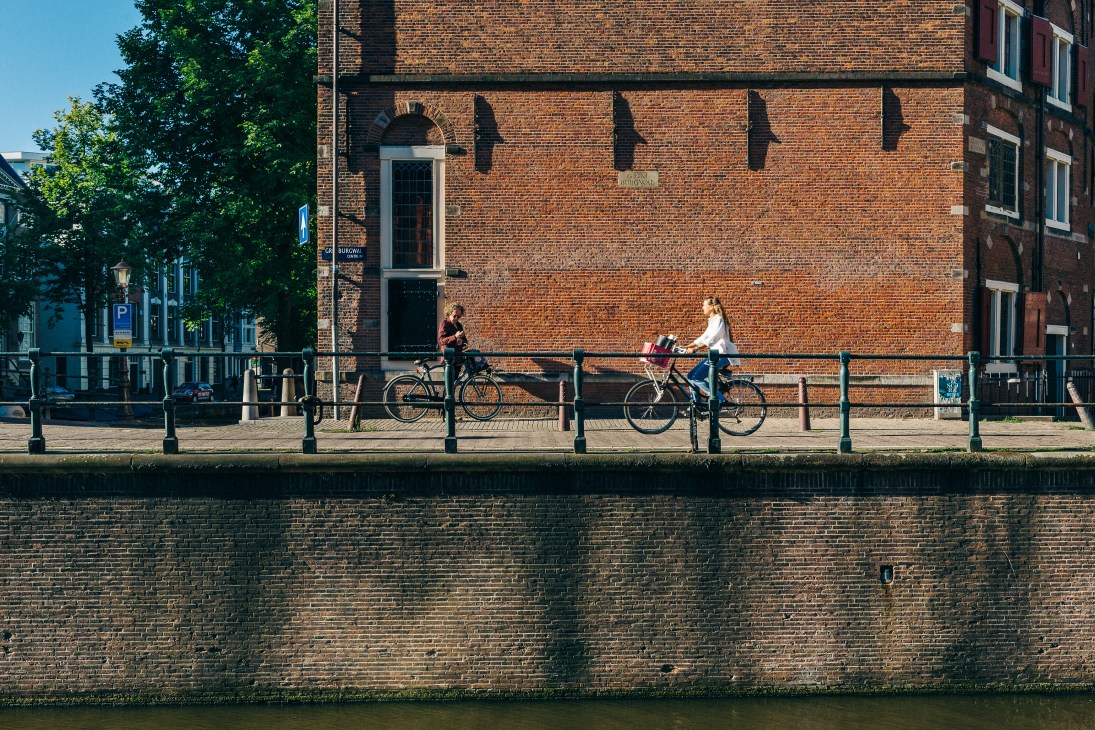 Cyclists ride along an Amsterdam canal beside a brick building on a sunny day.