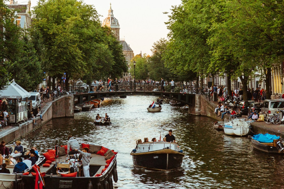 People relax on boats and canal banks in Amsterdam with a bridge and historic buildings in the background.