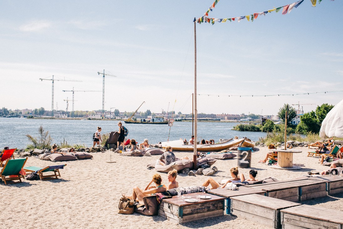 People relaxing on a sandy beach beside Amsterdam's waterfront with sailboats and industrial cranes in the background.