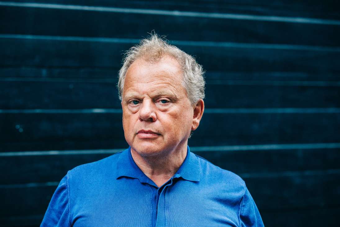 Bas Heijne, a middle-aged man with grey hair wearing a blue shirt, stands against a dark teal textured background.
