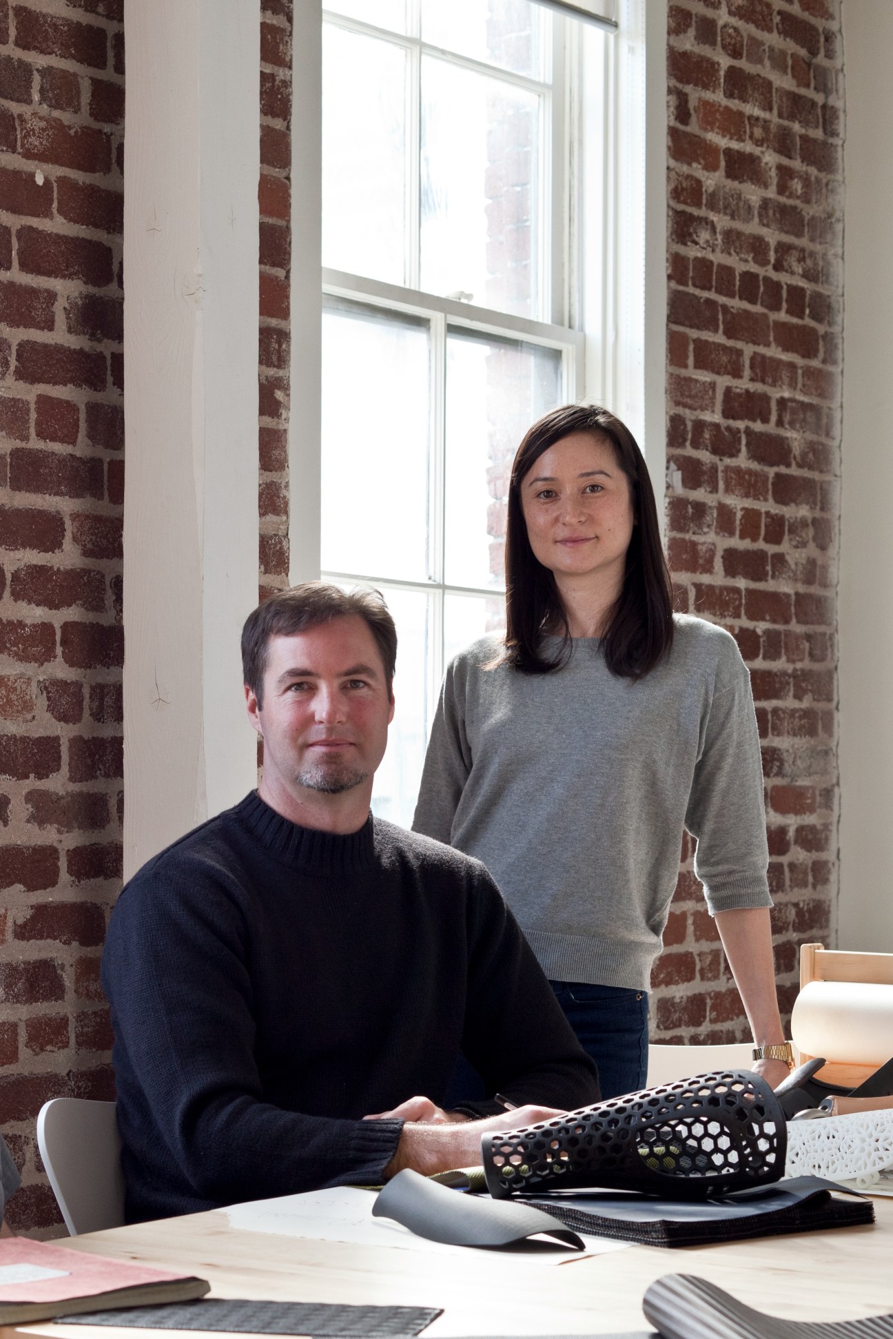 Two people sitting at a desk with fabric samples in front of them, against an exposed brick wall with large windows.