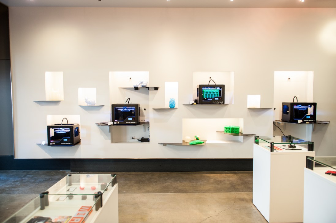 Makerbot 3D printers and printed objects displayed on white shelves in a retail store.