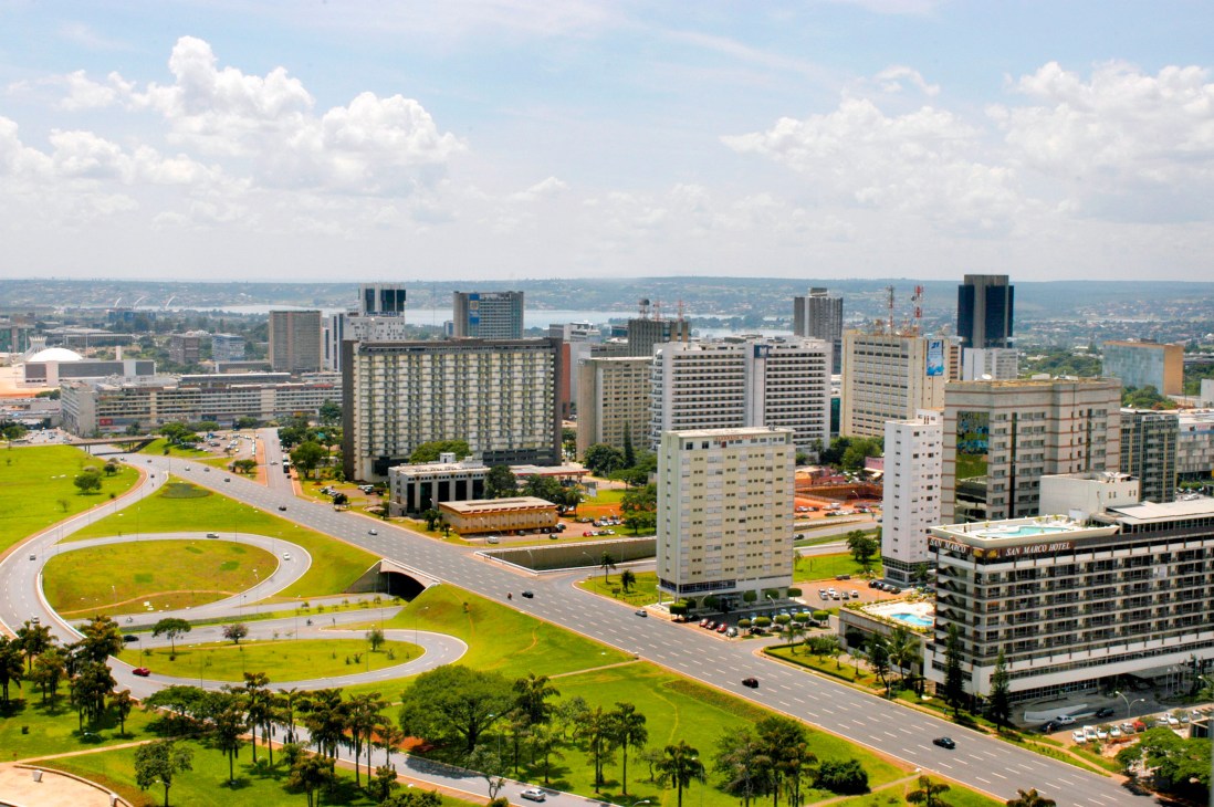 Aerial view of Brasilia's modernist architecture with green spaces, wide boulevards, and high-rise buildings under a blue sky.