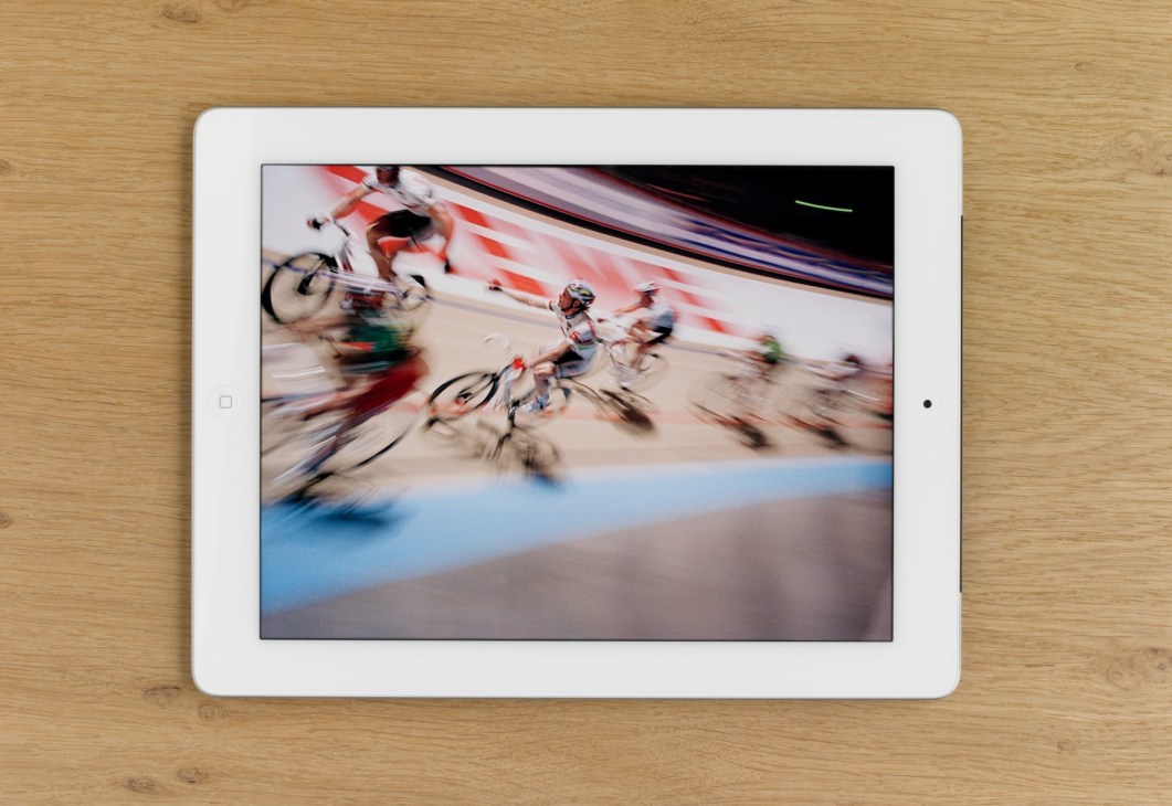 iPad displaying dynamic motion-blurred photograph of cyclists racing on an indoor velodrome track.