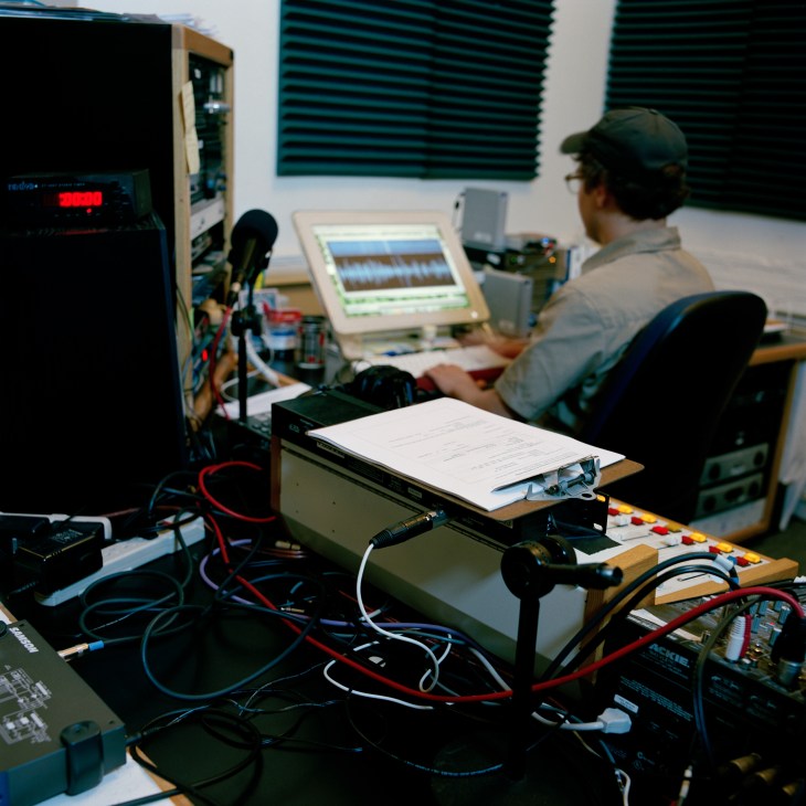 Radio station engineer working at broadcasting equipment with microphone, laptop displaying audio waveforms, and control panels.