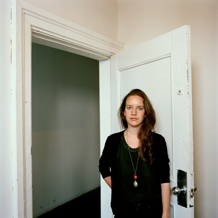 Beatrice Johnson, programme manager at AIR radio station, stands in a minimalist hallway wearing black clothing and a pendant necklace.