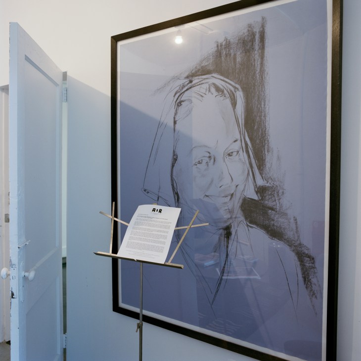 Framed portrait drawing of Alanna Heiss displayed on a wall with an AIR document on a stand.