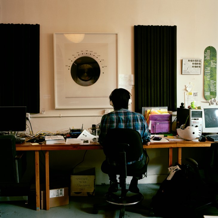 Radio broadcaster wearing headphones at desk in AIR studio with framed radio dial artwork on wall.