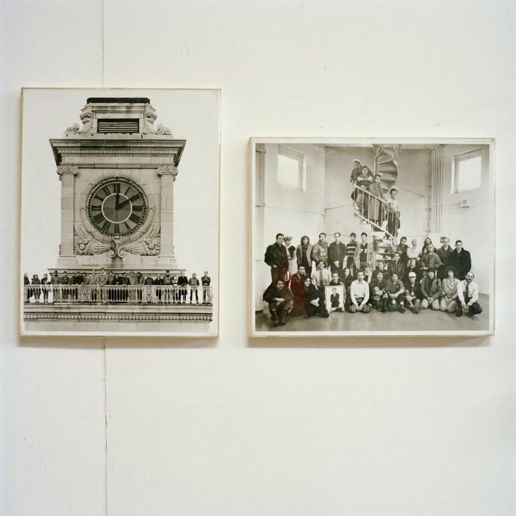 Two framed black and white photographs hanging on a white wall: one of an ornate clock tower with people on a balcony, and one of a large group of people posing together inside a building.