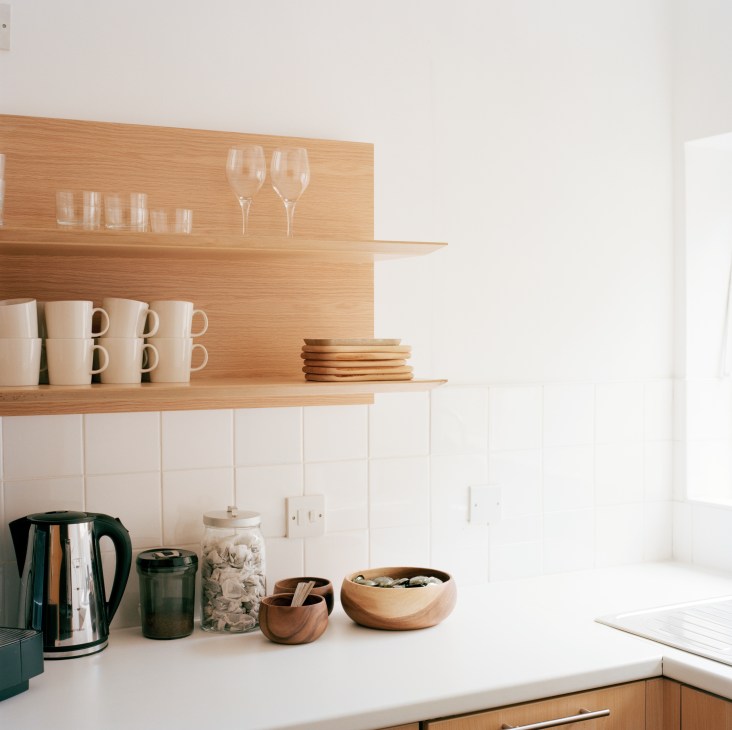 A minimalist kitchen area with wooden shelving displaying Iittala cups, glassware, and wooden accessories above a white countertop with a Nespresso machine.
