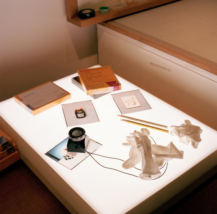 A illuminated lightbox display showing curated objects including books, sketches, watches, and white gloves arranged on a white surface.