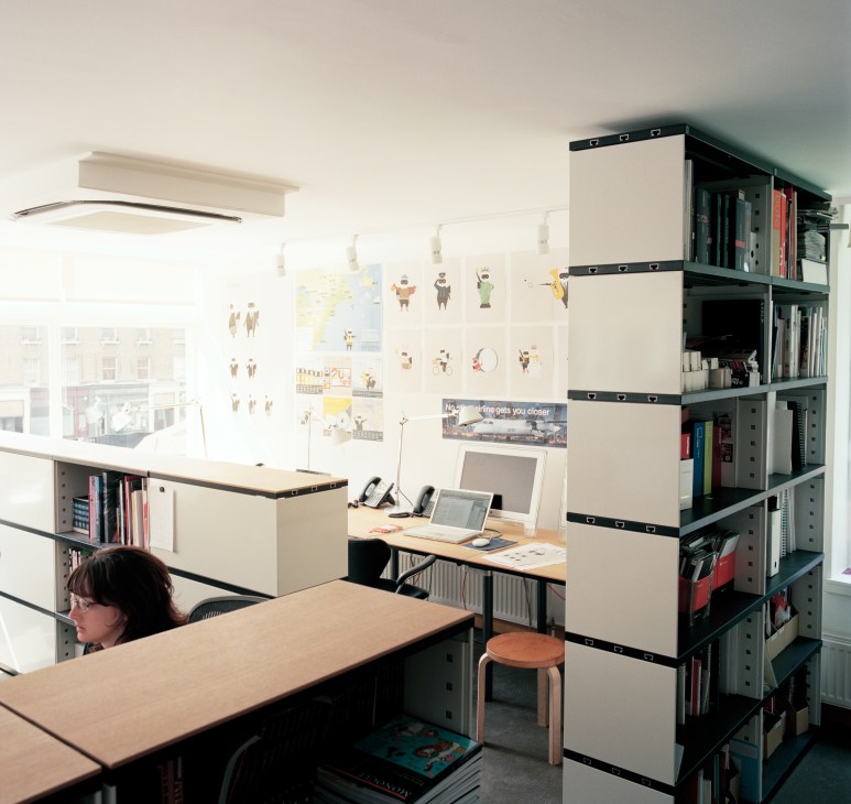 Modern office workspace divided by Novex shelving with oak desks, books, and an Artek stool visible.