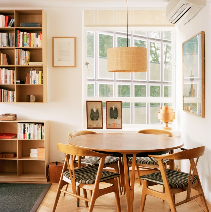Mid-century meeting room with round teak table, wooden chairs, bookshelves, and large windows overlooking green gardens.