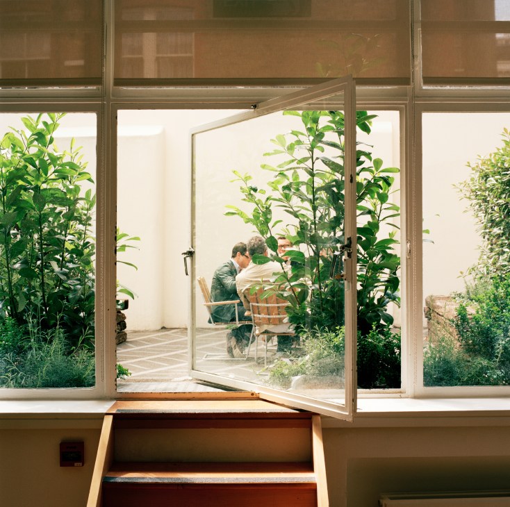 Two people dining at a wooden table in a lush green outdoor garden space viewed through open glass doors.
