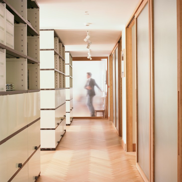 A modern corridor with oak parquet flooring, modular shelving units on the left, and wooden sliding doors on the right, with a blurred figure walking in the distance.