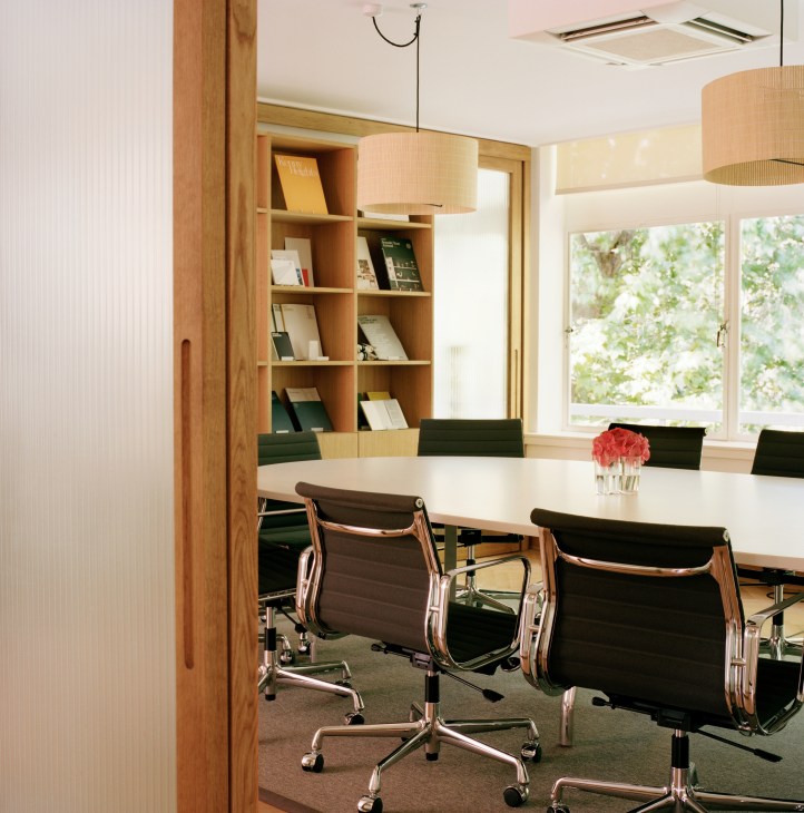 Modern meeting room with white conference table, black leather chairs, wooden shelving, and pendant lighting overlooking greenery.