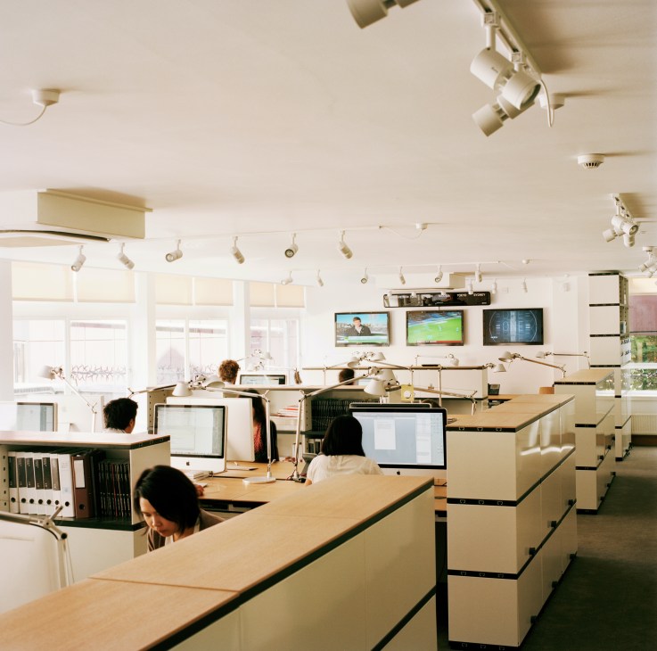 Monocle office with employees at oak desks with Novex shelving, Artemide task lamps and Panasonic televisions.