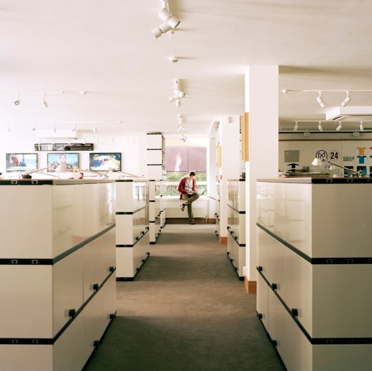 Open-plan office with white Novex shelving units dividing workstations, track lighting, and a person sitting at a desk in the center.