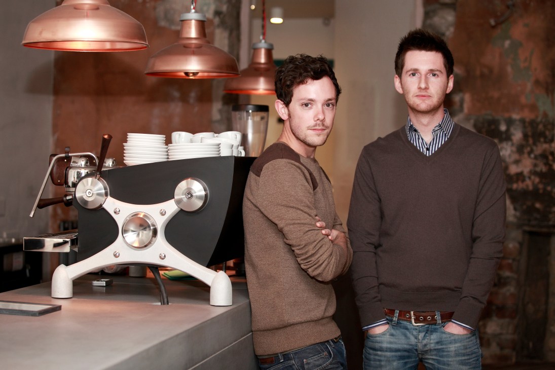 Two men stand beside a professional espresso machine with copper pendant lights above at Brew Lab coffee bar.