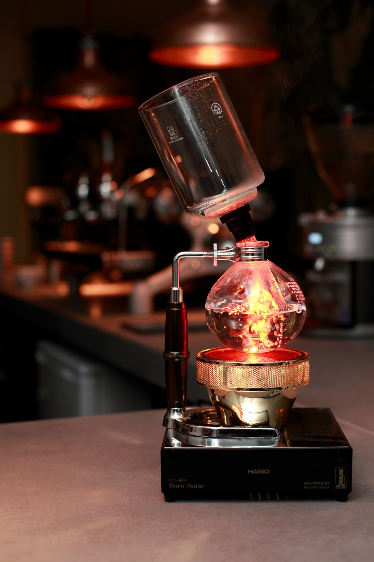 A Hario Beam Heater coffee siphon with glowing red bulb and glass vessel during brewing demonstration.