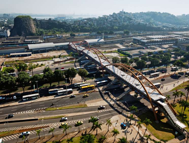 Aerial view of a curved steel footbridge and metro station spanning across Rio de Janeiro's urban infrastructure with rail yards and city beyond.