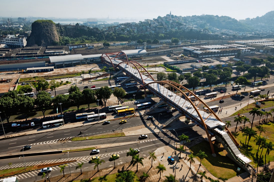 Aerial view of a curved steel footbridge and metro station spanning across Rio de Janeiro's urban infrastructure with rail yards and city beyond.