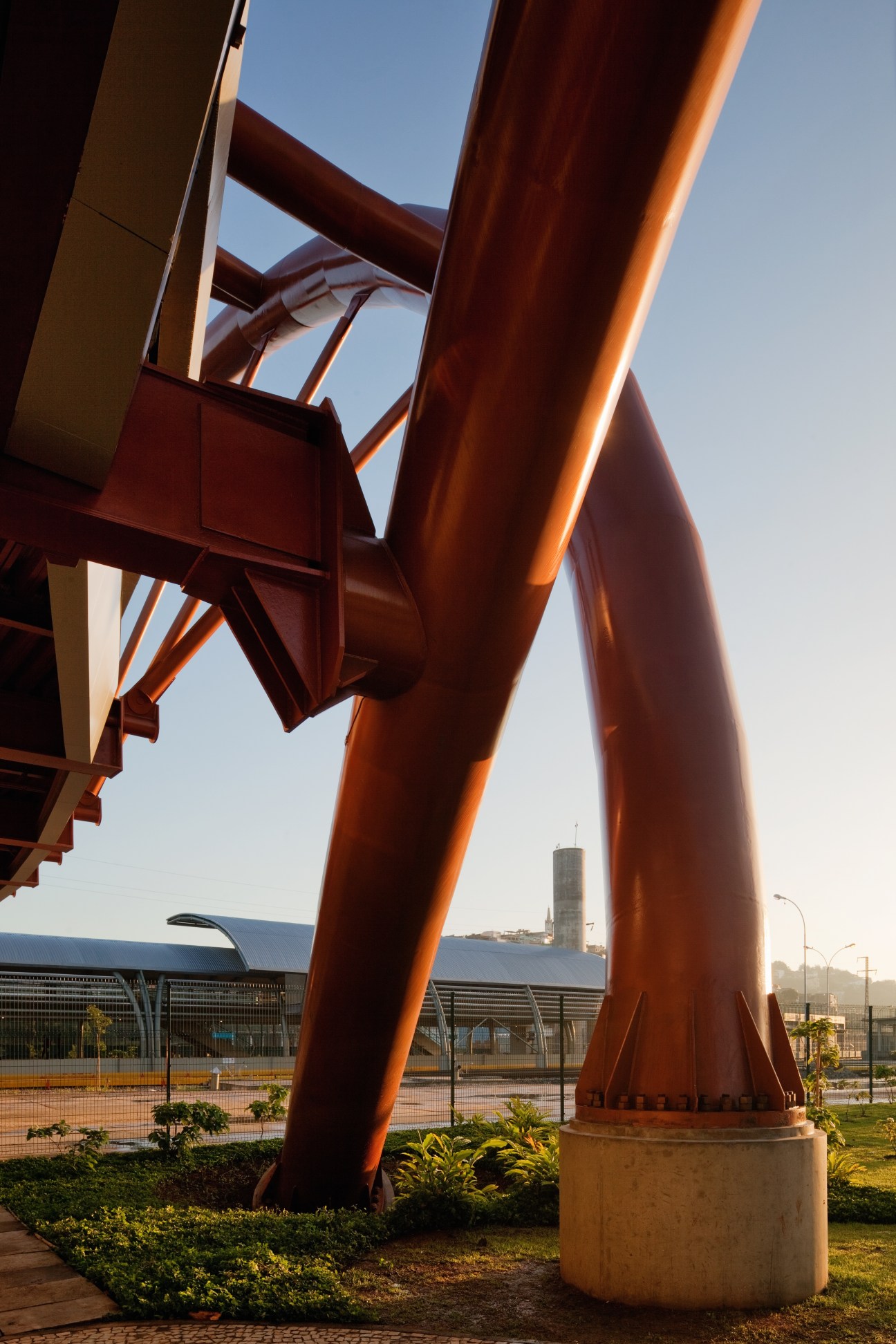 Red steel curved girders of a Rio metro station footbridge against clear blue sky.