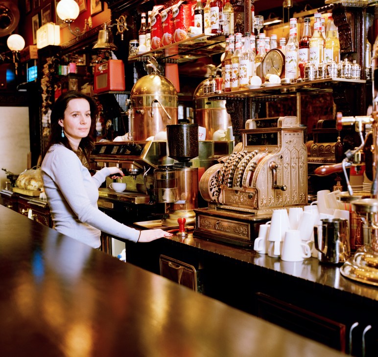 Barista Ricco stands behind the counter at The Bridge coffee bar in east London, with an ornate brass espresso machine and bottles of spirits displayed behind.