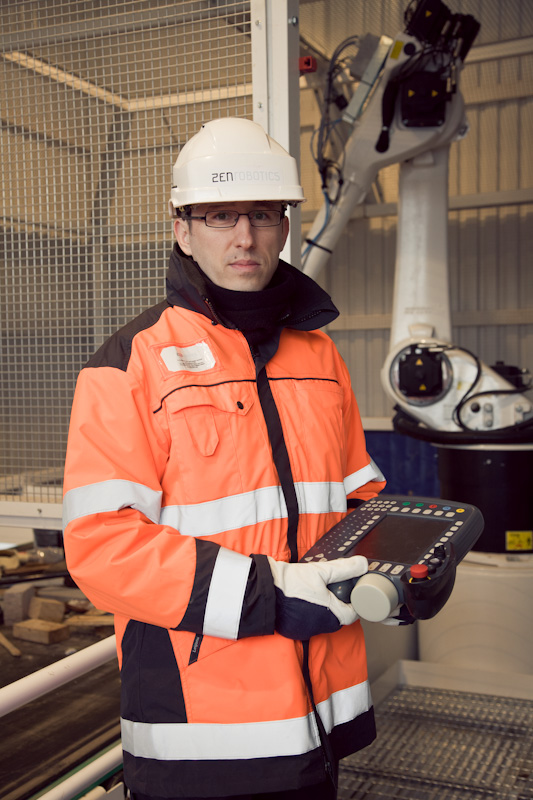 Man in safety helmet and orange reflective vest holding control device in front of robotic arm.