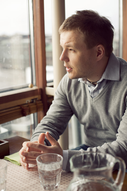 Man in grey sweater sitting by window, gesturing while speaking with glass of water nearby.