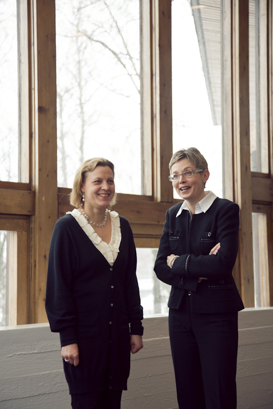 Two women standing by large windows, smiling at each other in an interior space.