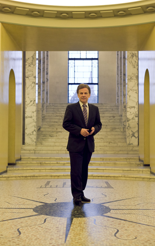 Finland's minister of economic affairs Mauri Pekkarinen standing in a modern government building corridor with yellow walls and marble columns.