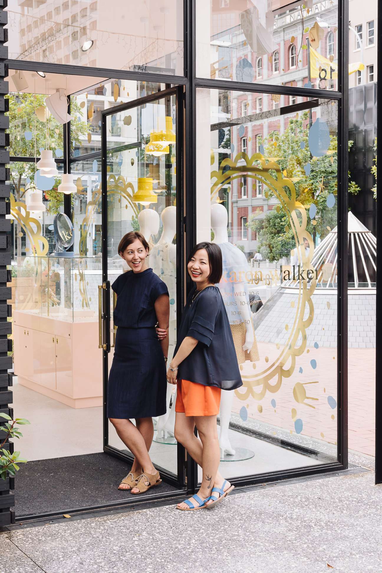 Two women smiling at the entrance of a modern retail storefront with large windows and decorative signage.