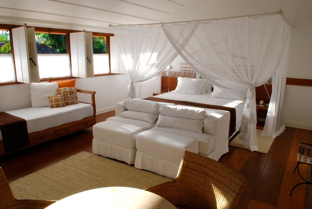 Bright bedroom with wooden frame canopy bed, white linens, cream seating area, and ocean views through windows.