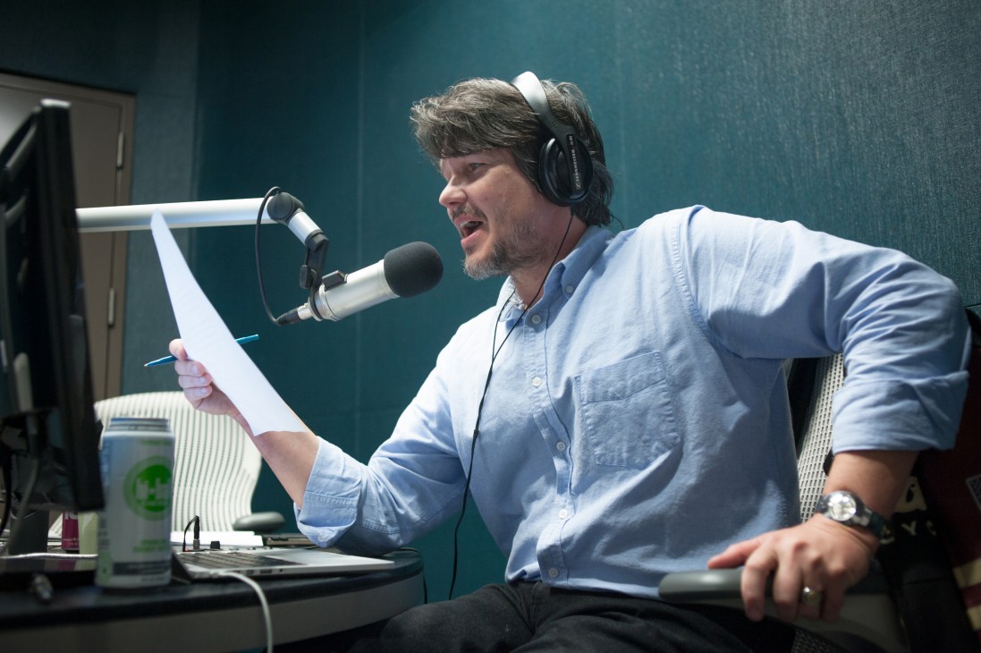 Man in blue shirt wearing headphones speaks into microphone at radio station studio desk.