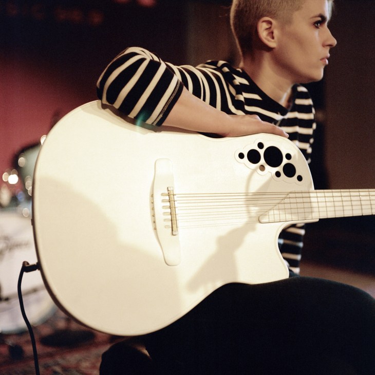 Musician Kaki King holds a white acoustic guitar while wearing a striped shirt in a radio studio.